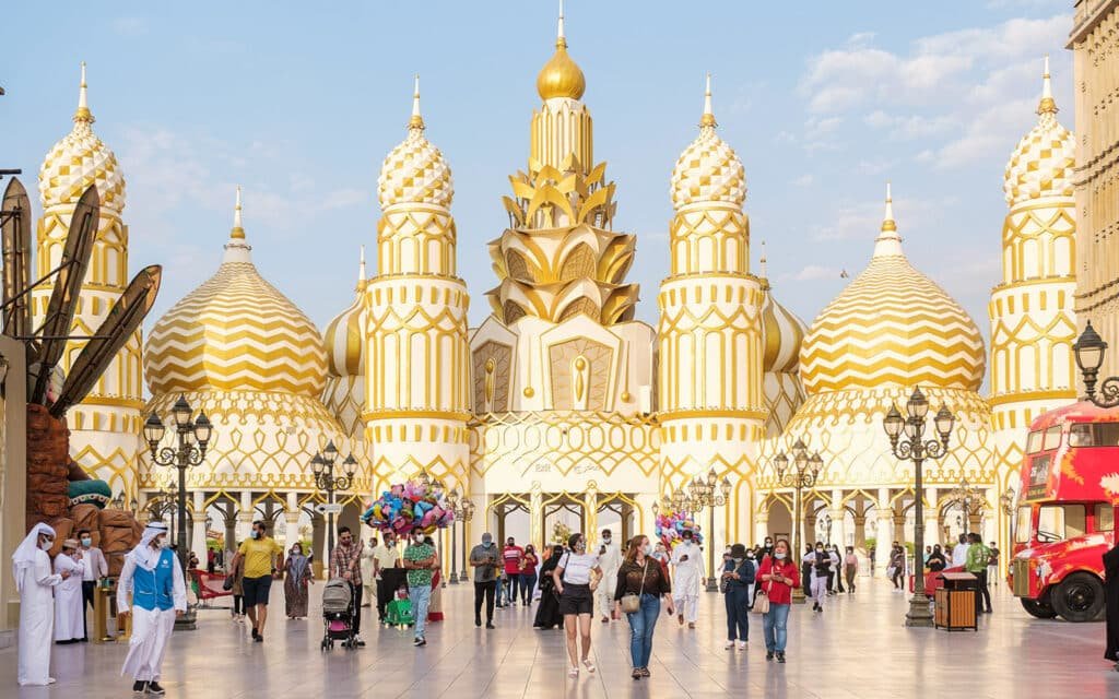 A crowd of people walks in front of a building with gold and white domes, resembling traditional Islamic architecture.