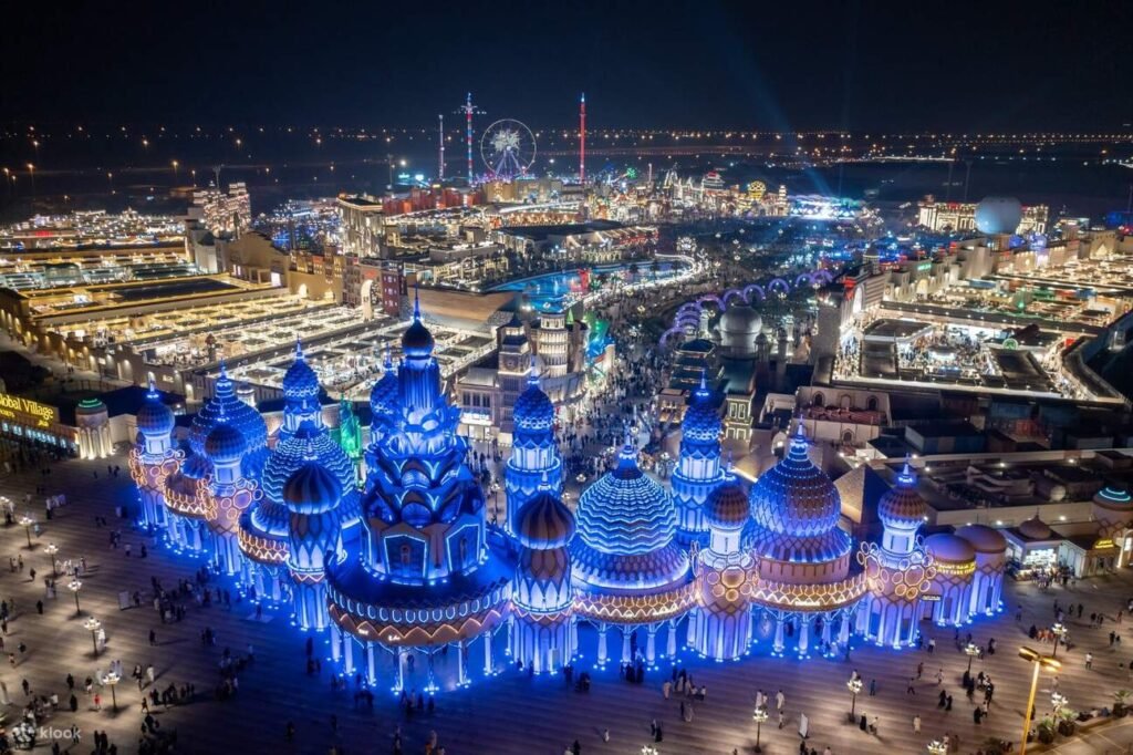 A vibrant night scene of an illuminated Global Village Dubai with domed buildings, colourful lights, and a distant Ferris wheel.