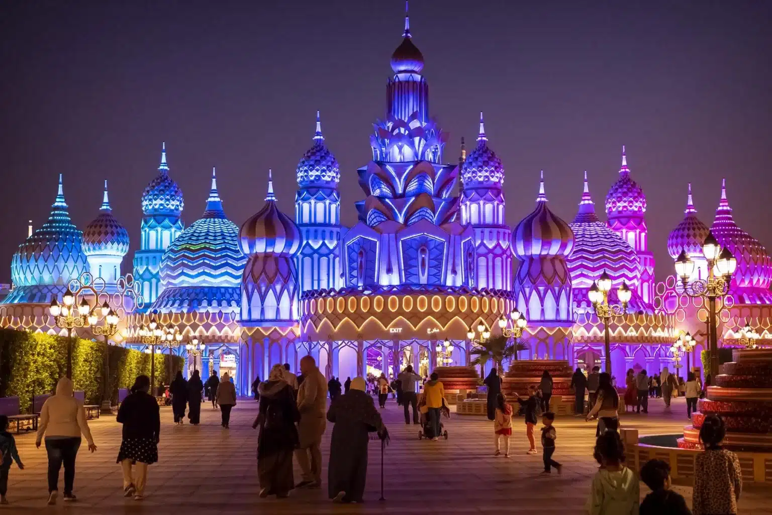 An ornate, brightly illuminated building with domes and spires at night, surrounded by walking visitors.