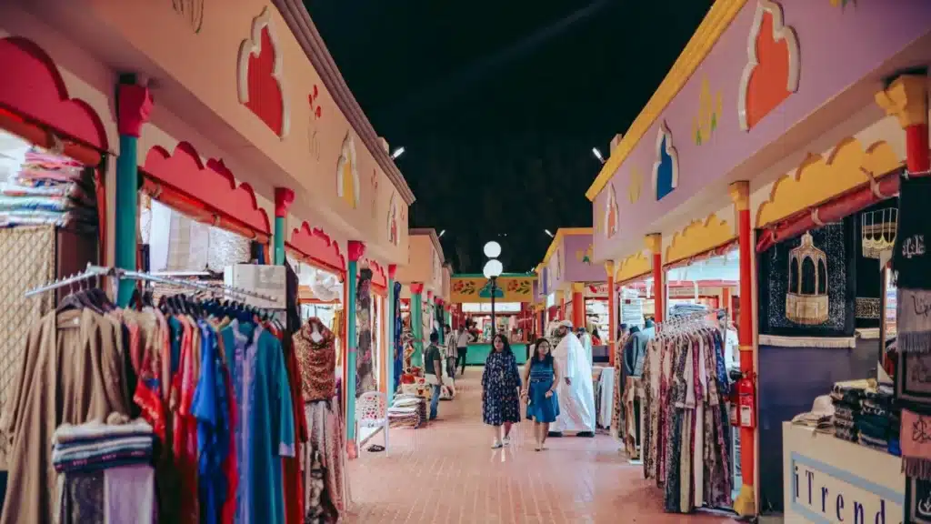 People walk through a vibrant market with colorful clothing displayed in open-fronted shops.