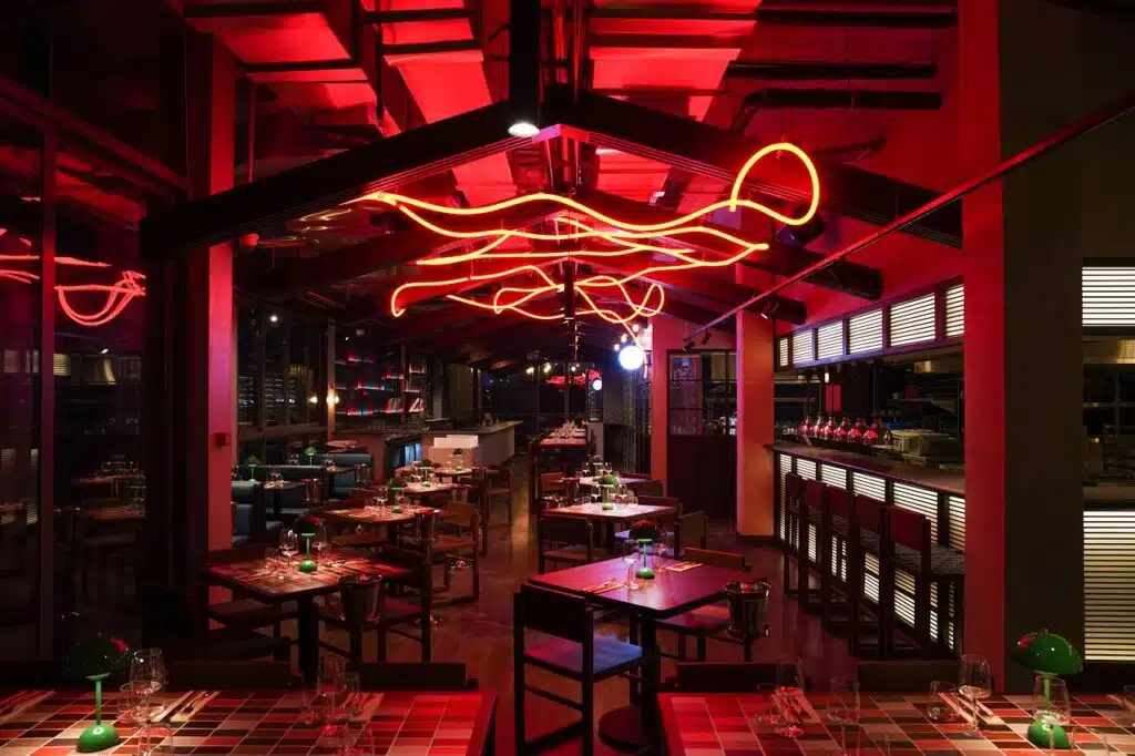 A dimly lit Hawkerboi restaurant interior with red neon light fixtures on the ceiling and tables set for dining.