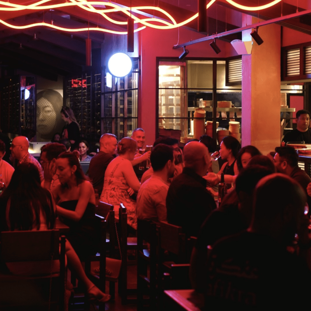 A dimly lit Hawkerboi restaurant with people seated at tables, illuminated by red neon lights, creating a lively atmosphere.