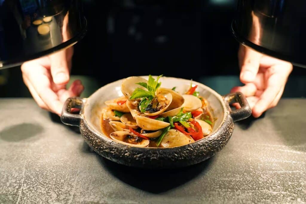 A bowl of clams garnished with herbs and vegetables is held by a person under warm lighting.