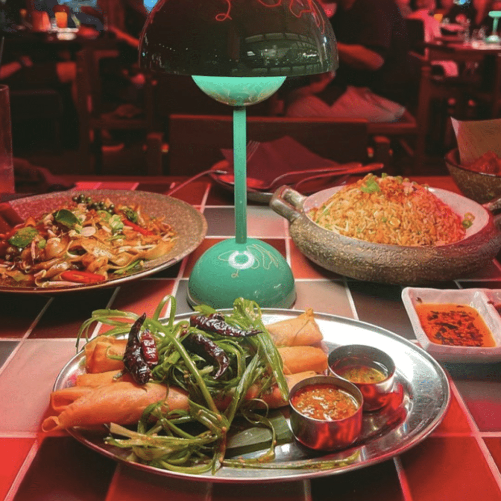A round Hawkerboi table with Asian dishes, including spring rolls with dipping sauces, noodles, and fried rice, under a green lamp.