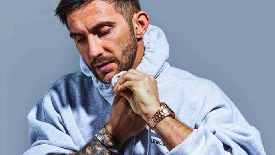 A man with a beard wearing a grey hoodie adjusts his collar, showcasing tattoos and a metallic watch.