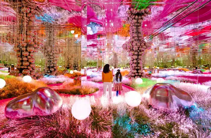 A vibrant, mirrored room filled with colorful lights, reflective surfaces, and two people walking among glowing orbs and plants.
