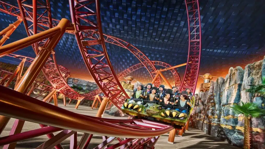 People enjoying a thrilling roller coaster ride with red tracks and a vibrant, colorful IMG Worlds of Adventure indoor setting.