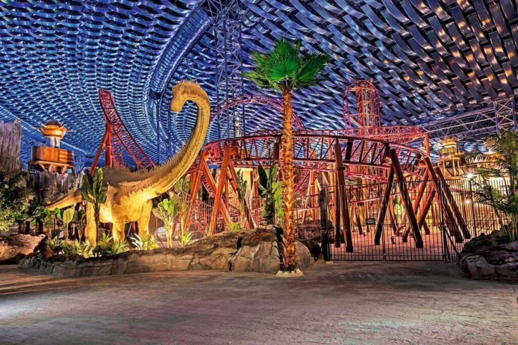 An indoor amusement park with a large dinosaur statue, roller coaster tracks, and a futuristic ceiling design.