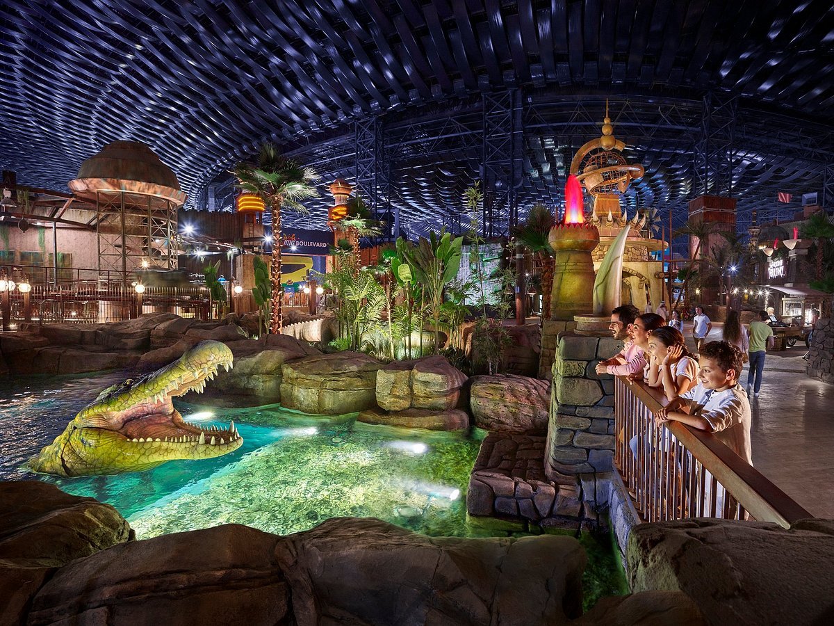 People observing an artificial crocodile in a themed indoor amusement park with tropical decor and dim lighting.