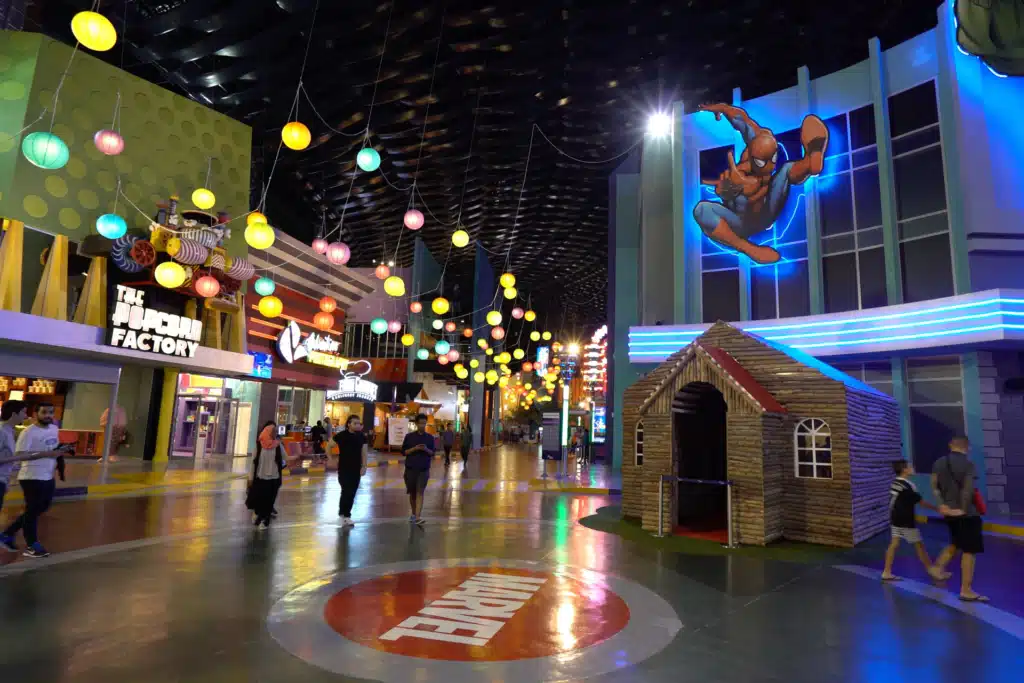 Indoor IMG Worlds of Adventure with colourful hanging lights, a Marvel theme logo on the floor, and people walking by a candy shop and playhouse.