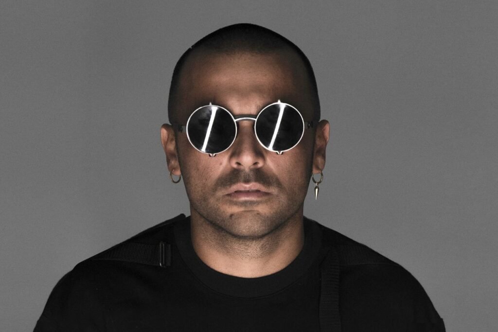 Ilario Alicante wearing round sunglasses, a dark shirt, and hoop earrings, looking directly at the camera against a gray background.