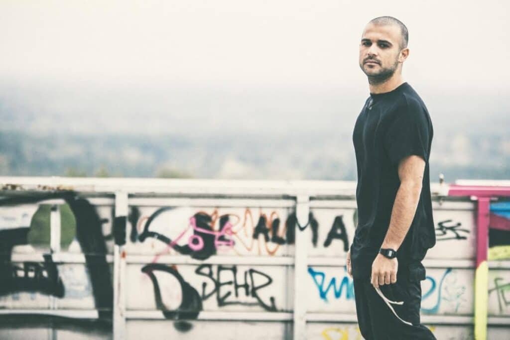 A person standing outside near a graffiti-covered wall, wearing a black outfit and a smartwatch.