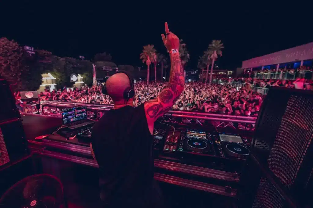 Ilario Alicante with tattoos, raises one arm while performing at an outdoor nighttime event, with a large crowd in the background.