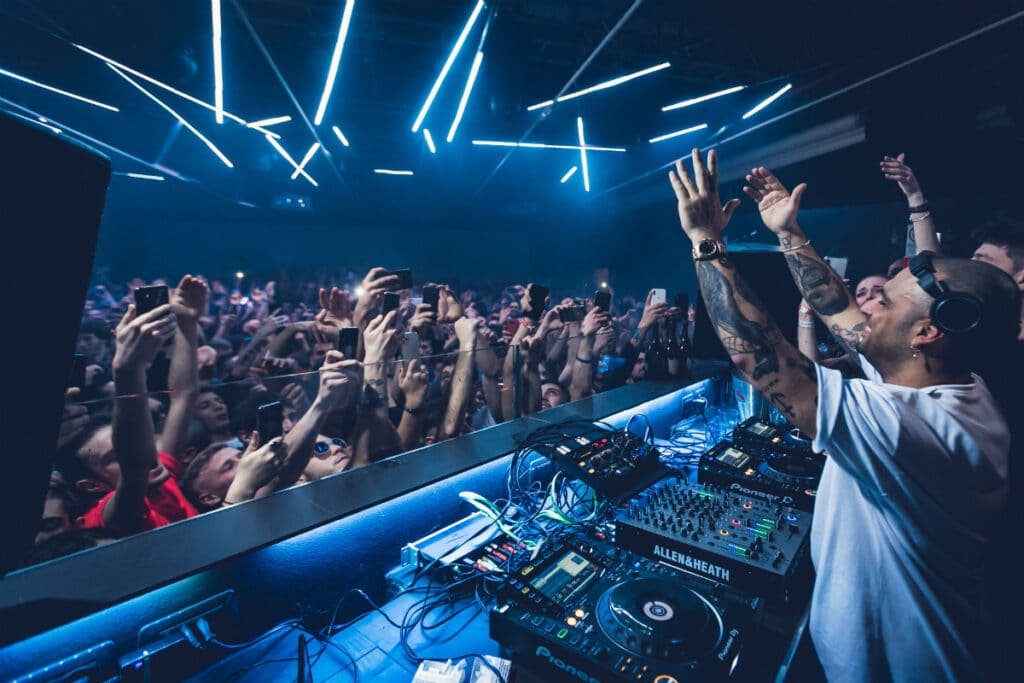 A DJ wearing headphones performs on stage, raising hands as a crowd cheers in a club with bright overhead lights.