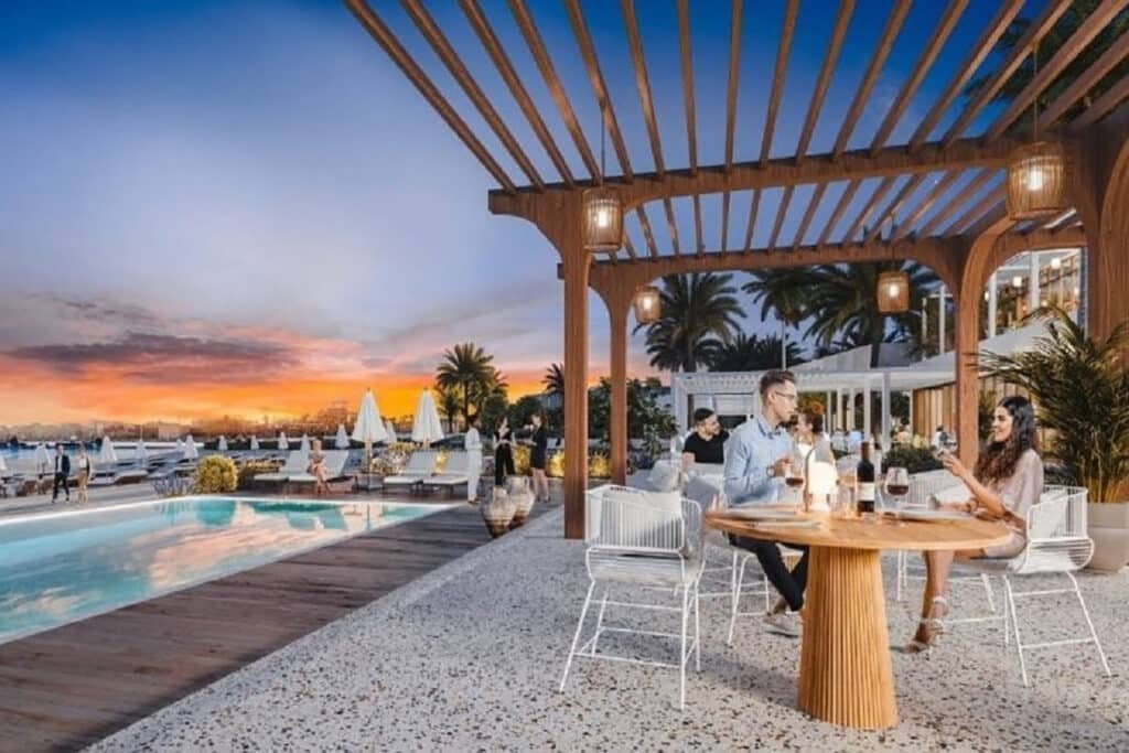 A couple sits at a patio table by a pool, enjoying drinks at sunset under a wooden pergola.