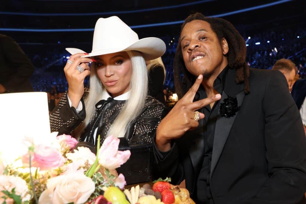 Beyonce in a white hat and sequined jacket, sits beside Jay-Z, making a peace sign, with flowers and fruit in the foreground.