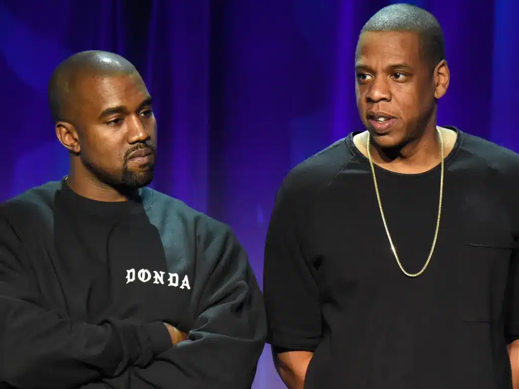 Two men on stage, one wearing a black "Donda" sweatshirt and the other a black T-shirt with a gold chain, against a blue background.