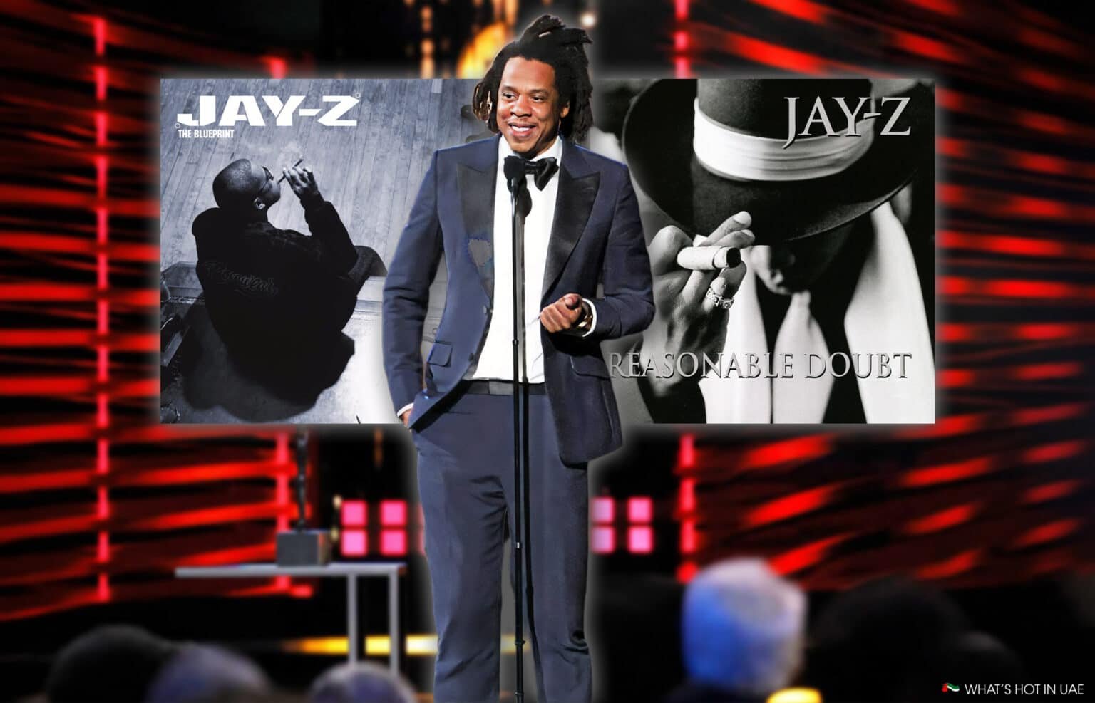 A person in a suit stands at a microphone on stage with two Jay-Z album covers, "The Blueprint" and "Reasonable Doubt," in the background.