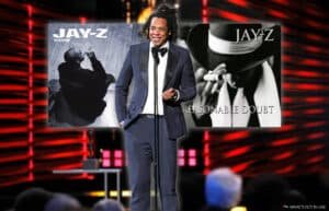 A person in a suit stands at a microphone on stage with two Jay-Z album covers, "The Blueprint" and "Reasonable Doubt," in the background.