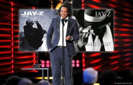 A person in a suit stands at a microphone on stage with two Jay-Z album covers, "The Blueprint" and "Reasonable Doubt," in the background.