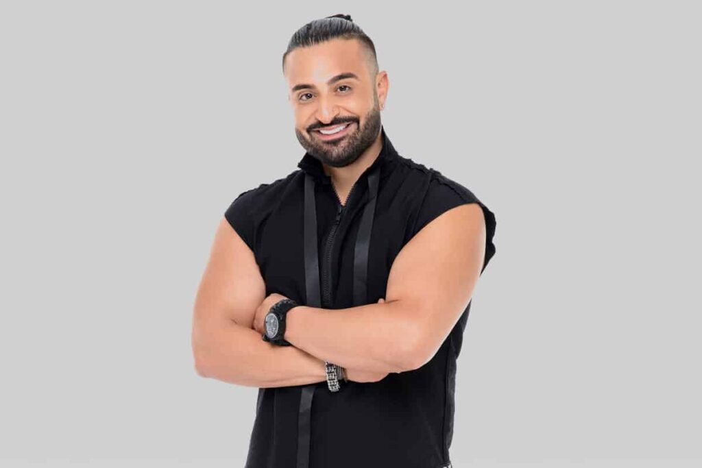 Man with a beard and ponytail smiling, wearing a sleeveless black shirt and a watch, standing against a gray background.