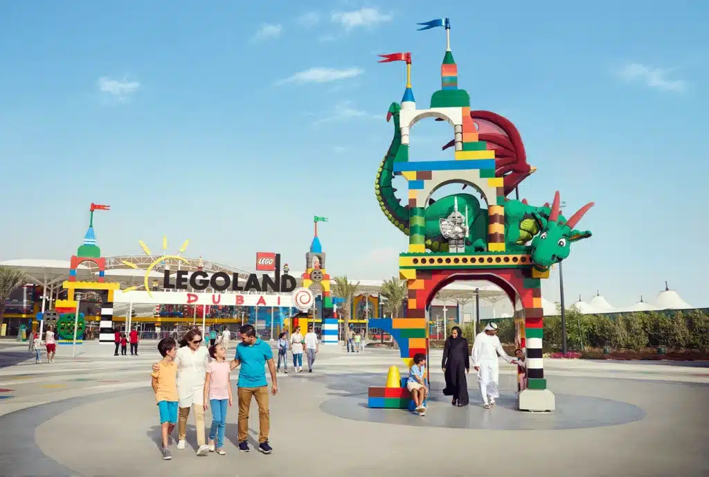A group of people walk near a colorful LEGO dragon arch at Legoland Dubai on a sunny day.