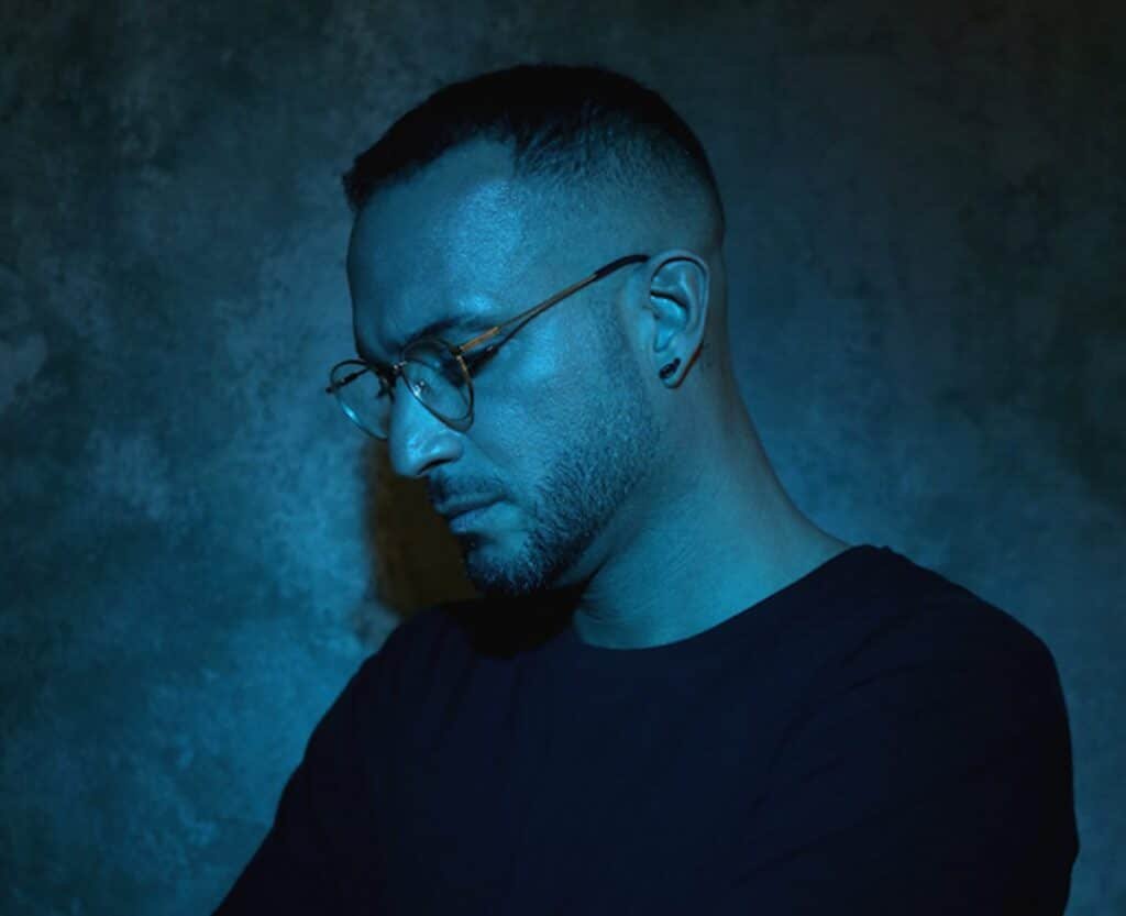 A man with glasses and a beard is illuminated by blue lighting against a textured, dark background.