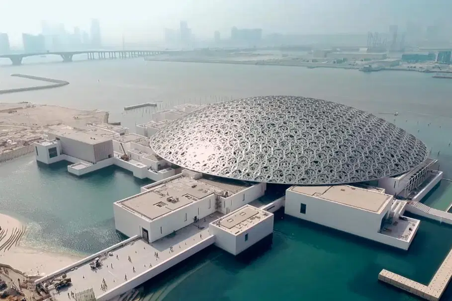 The Best Free Things Worth Doing in the UAE: Aerial view of the louvre with intricate patterns, surrounded by water and connected buildings in a coastal cityscape.