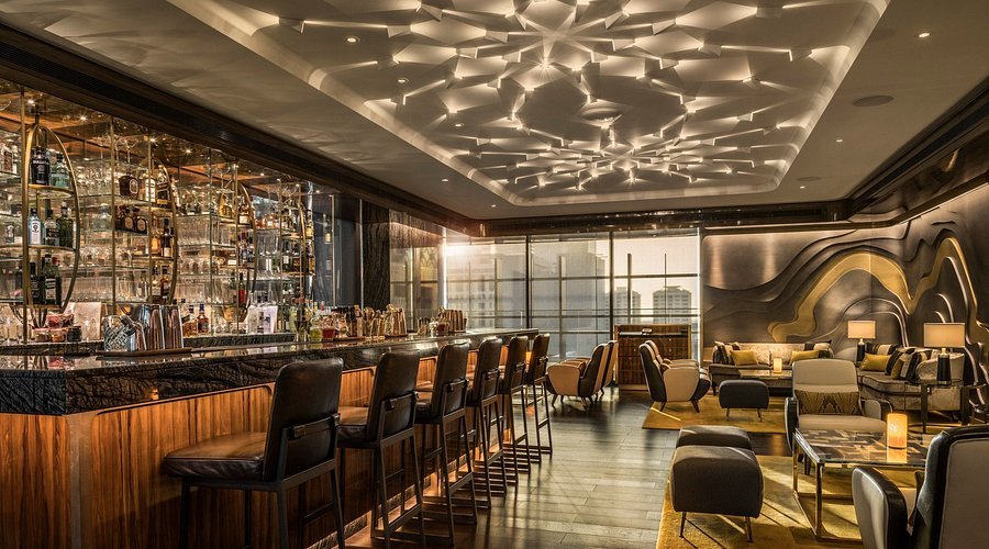 Luxurious bar with modern decor, high stools at the counter, glass shelves with bottles, and elegant seating area with city view.