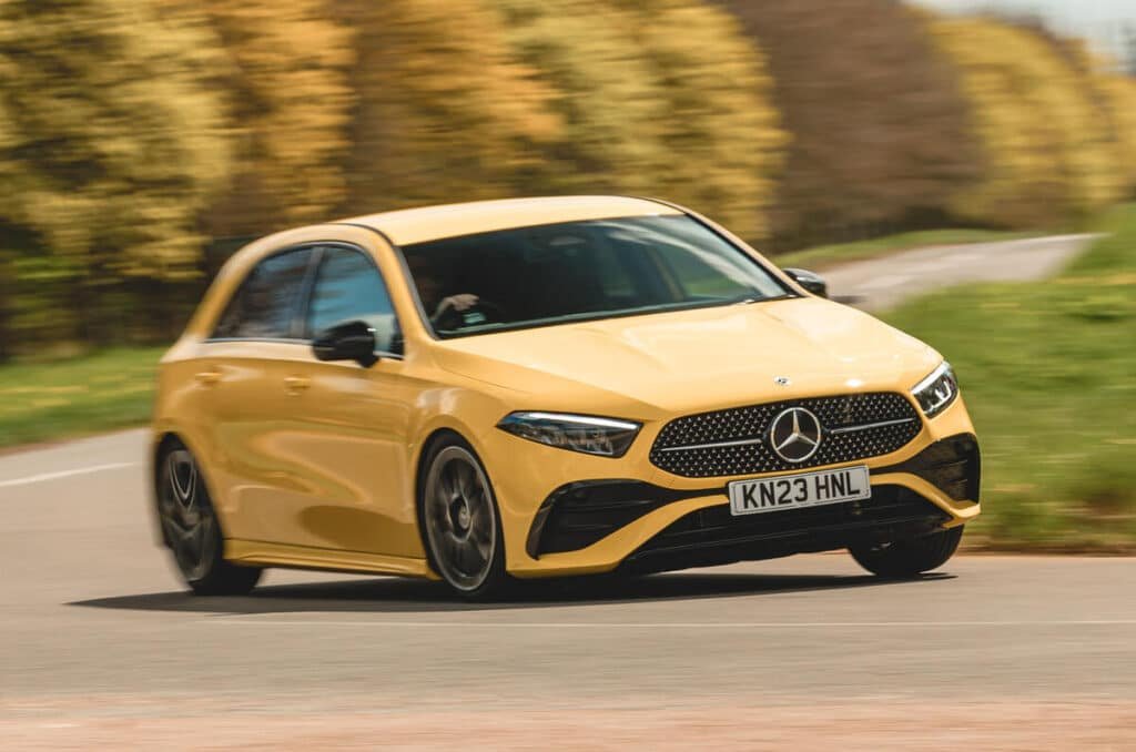 A yellow Mercedes-Benz A-Class 2028 hatchback driving on a curved road, surrounded by blurred greenery.