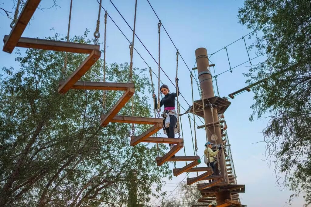 The Best Free Things Worth Doing in the UAE: A person is balancing on a suspended wooden obstacle course high above the ground, surrounded by trees and a clear sky.