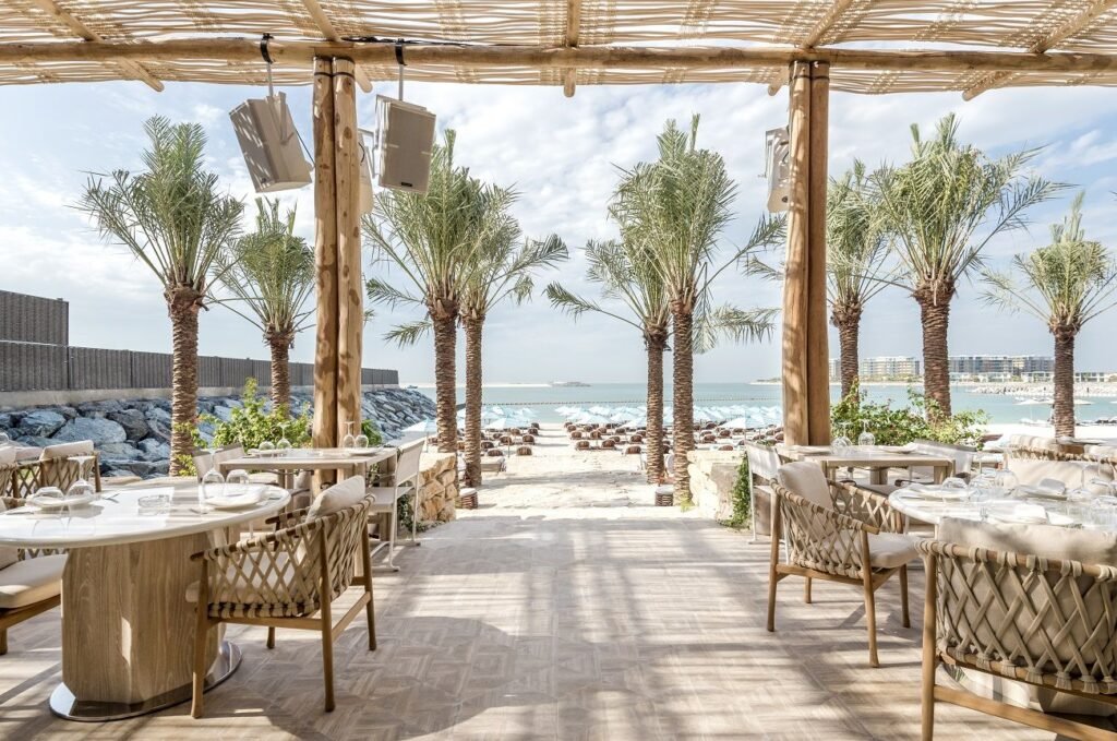 Outdoor Nammos Dubai beachside restaurant with palm trees, wooden pergola, and view of the ocean and lounge chairs on the sand.