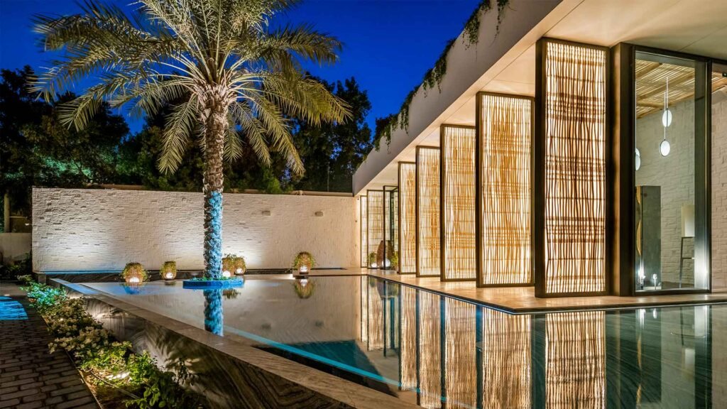 Modern house at night with large illuminated windows, palm tree, and a reflective swimming pool.
