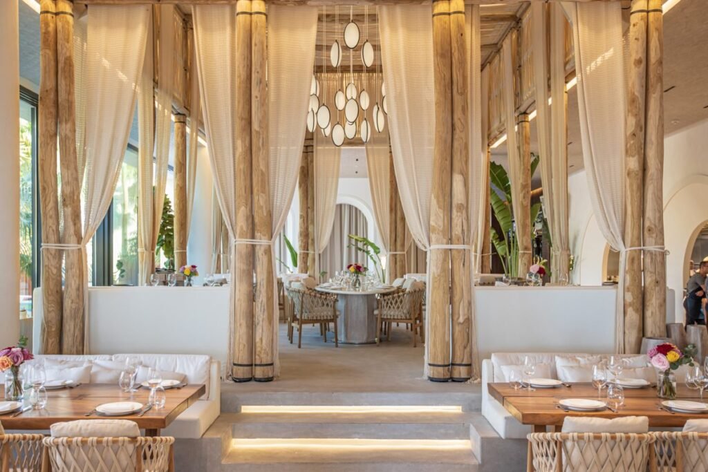 Elegant Nammos Dubai restaurant interior with wooden columns, white curtains, and tables set with white plates and floral centerpieces.