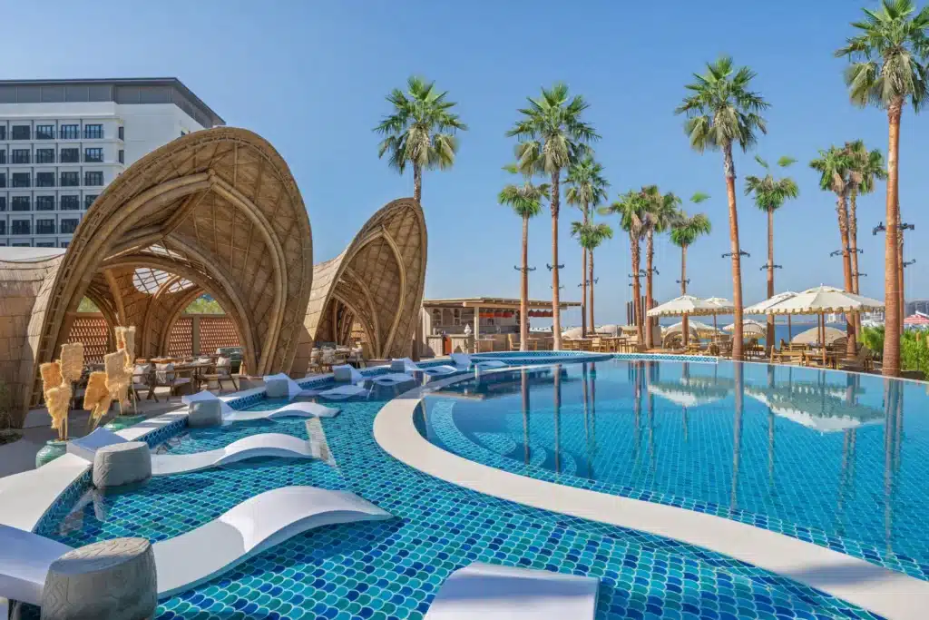 A luxurious poolside area with modern lounge chairs, cabana-style structures, and tall palm trees under a clear blue sky.