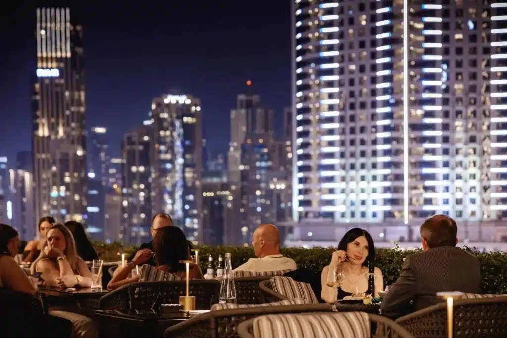People dining outdoors at night on a terrace with a cityscape of illuminated skyscrapers in the background.