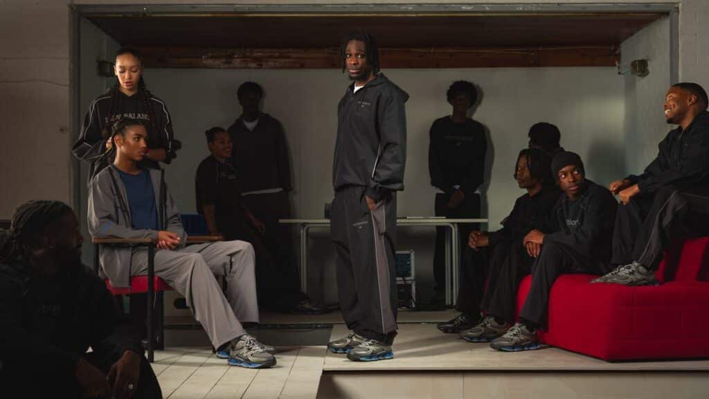 Several people are wearing casual streetwear, with one person standing while others sit on red seating in a dimly lit room.
