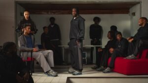 Several people are wearing casual streetwear, with one person standing while others sit on red seating in a dimly lit room.