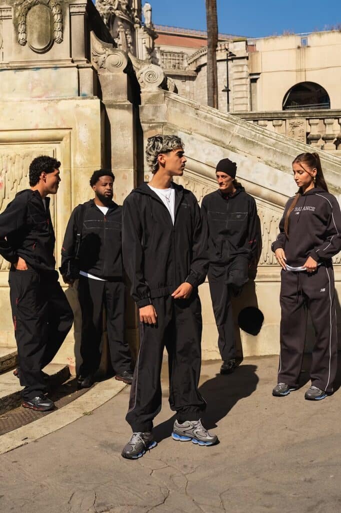 Five people wearing casual black tracksuits stand together on a sunlit stone path near ornate stonework.