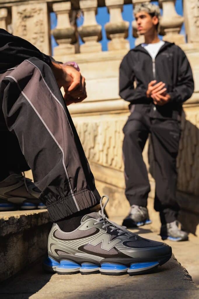 Two people in athletic wear, one seated showing a close-up of a New Balance ABZORB 2000 sneaker with blue accents, and the other standing in the background.