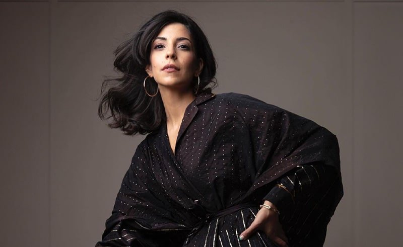 A person with long dark hair, wearing a stylish black outfit, poses confidently.