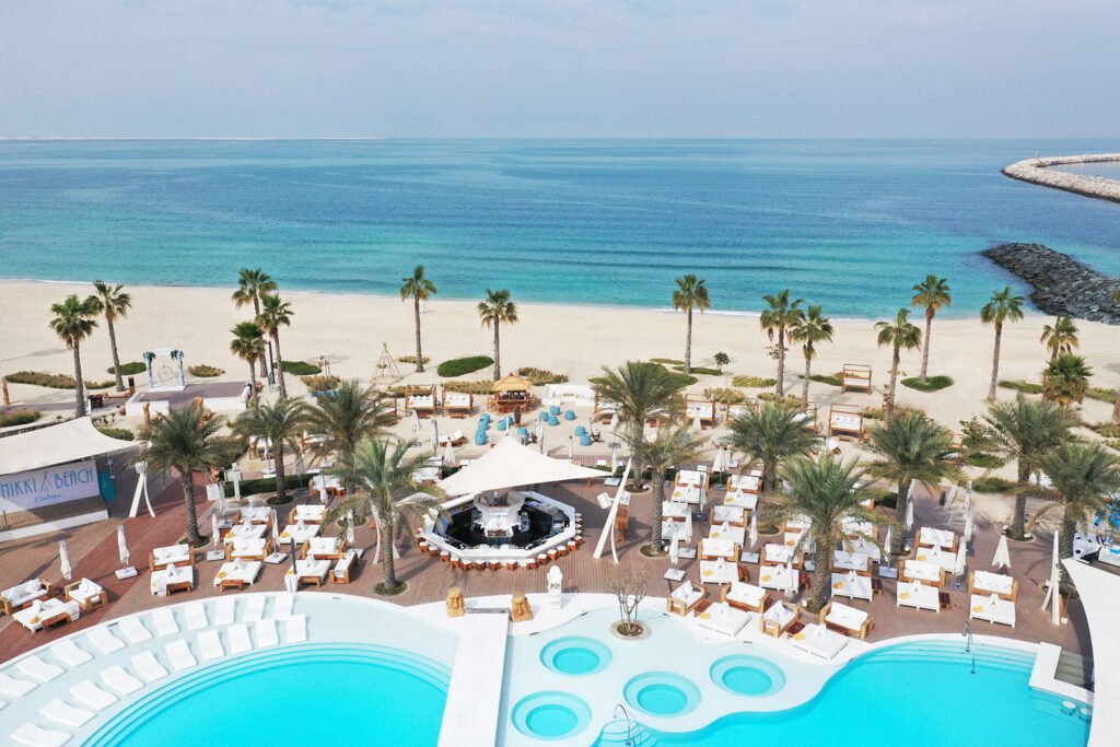 Nikki Beach resort with palm trees, swimming pools, and lounge areas overlooking a calm blue ocean.