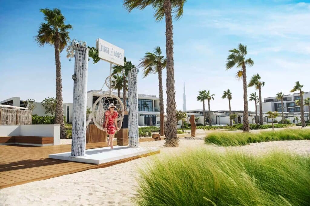 A person stands on a swing under a decorated archway on a sandy beachfront with palm trees and modern buildings nearby.
