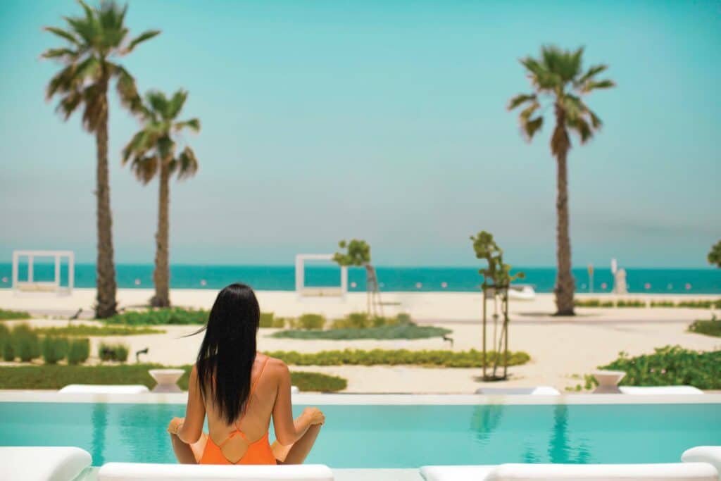 Woman in an orange swimsuit sitting by the Nikki Beach pool, facing a beach with palm trees and clear blue sky.