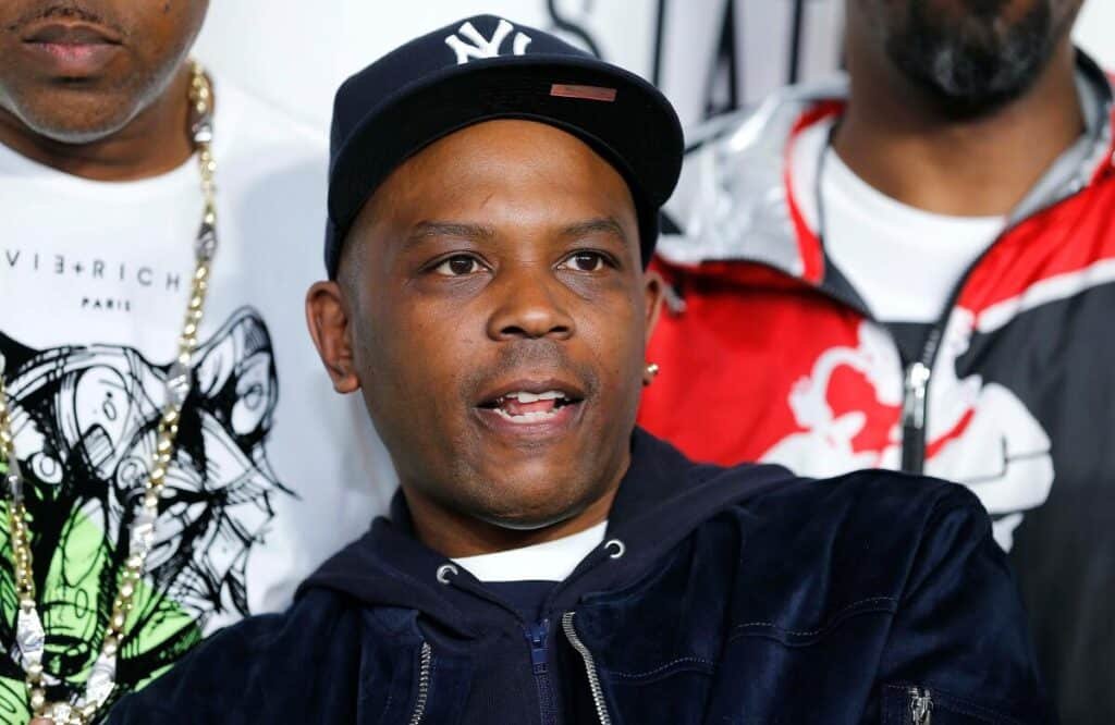 Three men are gathered closely, with the central figure wearing a navy cap and jacket, standing in front of a wall with logos.