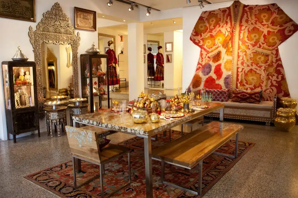 A dining room with a long table adorned with metallic serving dishes and goblets, surrounded by decorative cabinets and vibrant textiles.