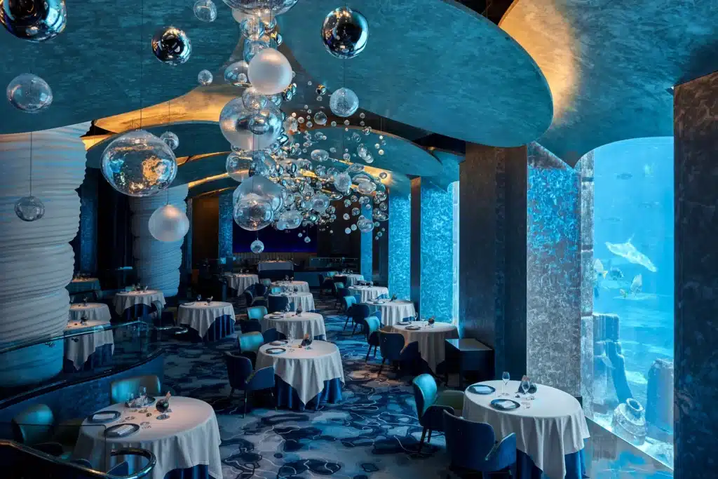 An elegant underwater-themed restaurant with round tables, blue accents, and large aquarium windows displaying marine life.