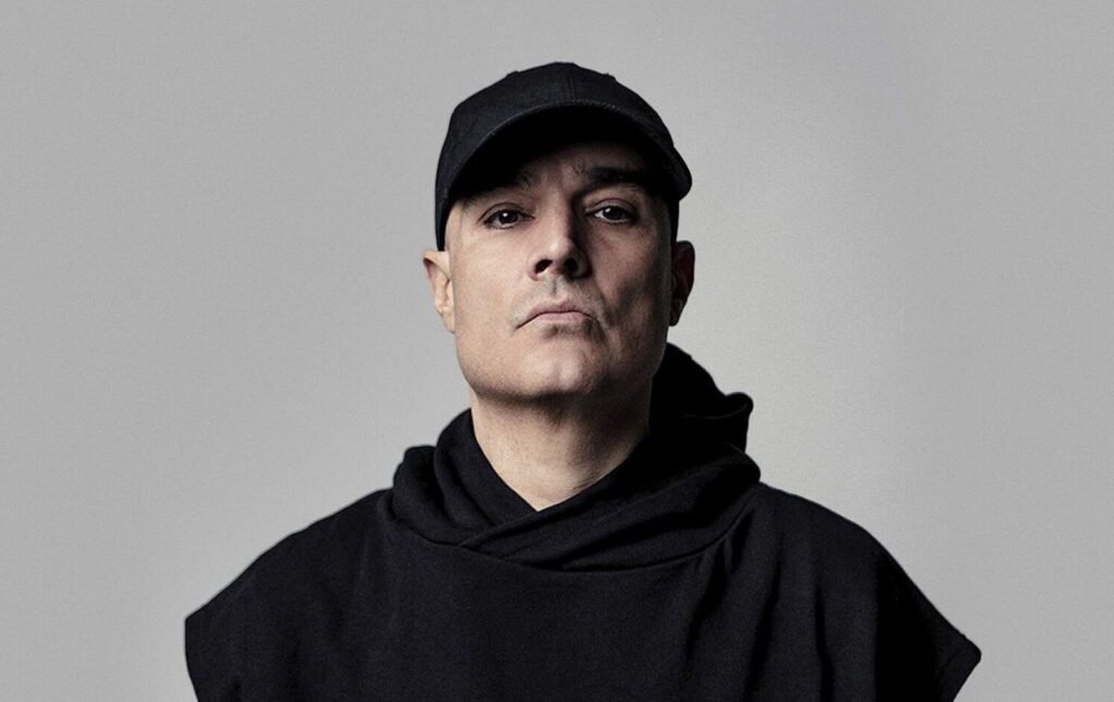 A person wearing a black cap and black clothing, looking directly at the camera against a plain background.