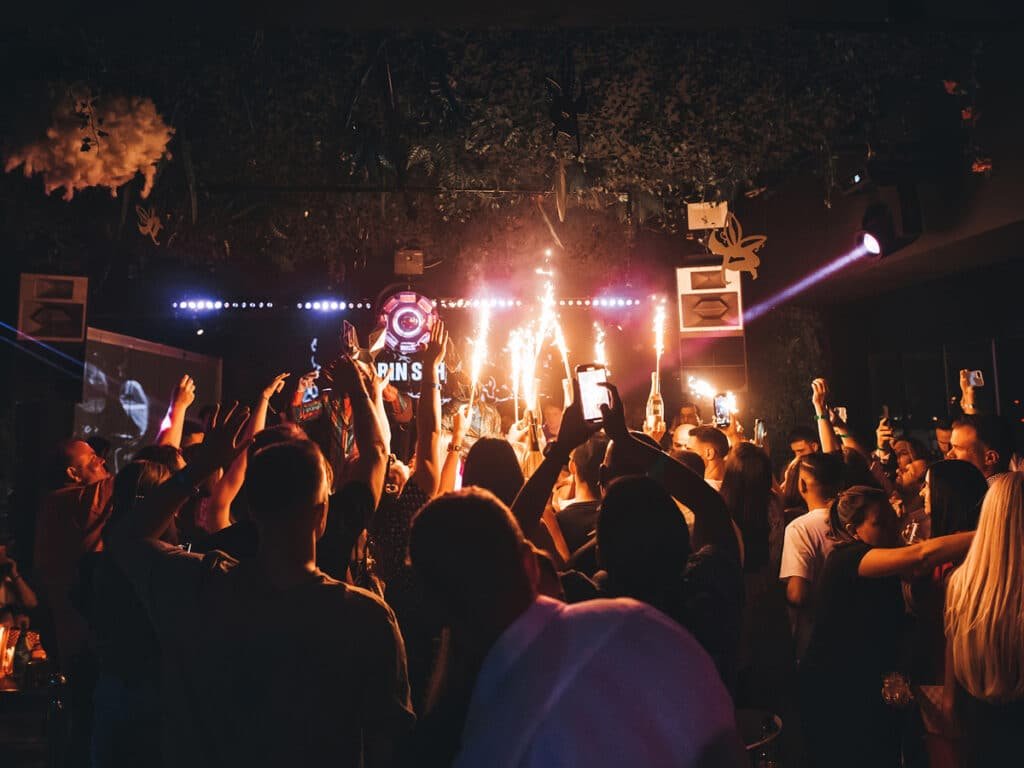 A lively crowd at a nightclub, holding up phones and drinks as sparklers light up the room.