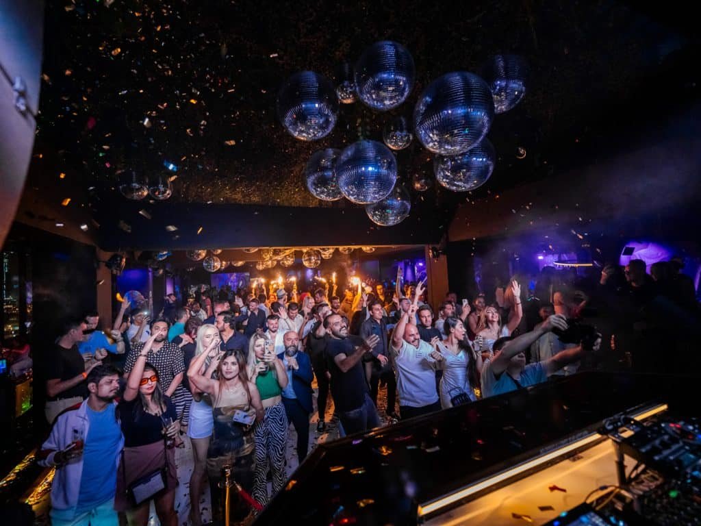 Crowd dancing energetically in a dimly lit nightclub with disco balls and confetti falling from the ceiling.